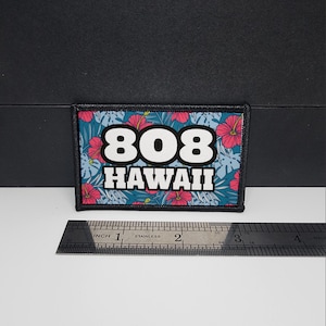 Könnte beinhalten: Rechteckiger Aufnäher mit dem Text "808 HAWAII" in Weiß, vor einem tealfarbenen Hintergrund mit rosa Hibiskusblüten und grünen Blättern. Der Aufnäher ist etwa 7,6 cm breit.