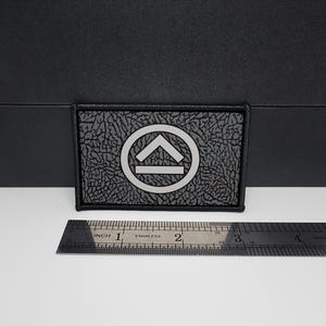 May include: A rectangular black patch with a textured surface and a silver emblem. The emblem features a circle with an upward-pointing arrow and a horizontal line. A stainless steel ruler is placed below the patch, measuring in inches.
