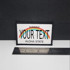 以下が含まれることがあります： 白い背景、黒い縁取り、テキスト「HAWAII YOUR TEXT ALOHA STATE」の長方形パッチ。虹がテキストの上にあります。定規は下に配置され、センチメートル単位の測定値を示しています。
