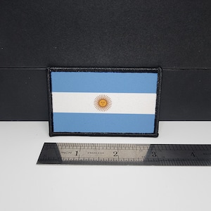 May include: Rectangular patch featuring the flag of Argentina. The flag has three horizontal stripes: two light blue stripes sandwiching a white stripe. The center of the white stripe features a golden sun with a face. The patch is bordered with black stitching.
