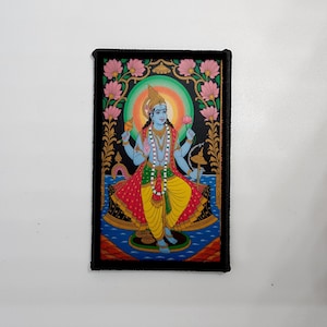 Könnte beinhalten: Ein rechteckiger Aufnäher mit einer lebendigen Illustration einer Gottheit mit blauer Haut, geschmückt mit einer goldenen Krone und Schmuck. Die Figur sitzt, umgeben von rosa Lotusblüten und einem grünen und orangefarbenen Heiligenschein. Der Aufnäher hat einen schwarzen Rand.