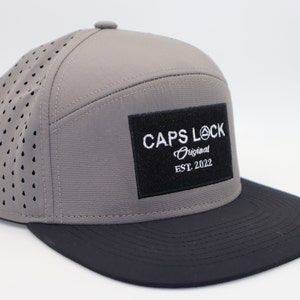 Puede incluir: Gorra de béisbol gris y negra con el texto "CAPS LOCK Original EST. 2022" en un parche negro. La gorra presenta un diseño perforado en los paneles laterales y una visera plana.