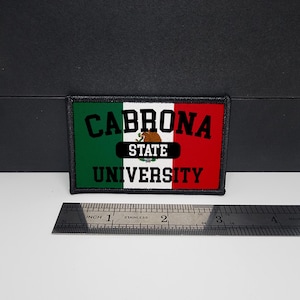 May include: Rectangular patch with the text "CABRONA UNIVERSITY" in white letters over a green, white, and red background, resembling a flag. The patch includes a brown bear graphic and the words "STATE" above "UNIVERSITY". A stainless steel ruler is placed below the patch.