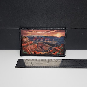 Könnte beinhalten: Rechteckiger Aufnäher mit einer malerischen Illustration des Grand Canyon bei Sonnenuntergang. Der Aufnäher hat einen schwarzen Rand und den Text "GRAND CANYON" oben. Das Bild zeigt die geschichteten Felsformationen des Canyons in Rot-, Orange- und Blautönen.