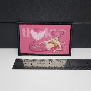 May include: A rectangular pink patch with a black border. The patch features a mermaid illustration and the word "thank" in white. The mermaid has pink hair and a pink and purple tail. A ruler is below the patch.