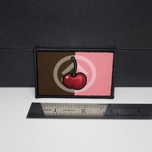 Peut inclure: Écusson rectangulaire à bordure noire, divisé en sections marron et rose. Une cerise rouge avec une tige verte est centrée sur l'écusson. Une règle en acier inoxydable est en dessous, avec des mesures en cm.