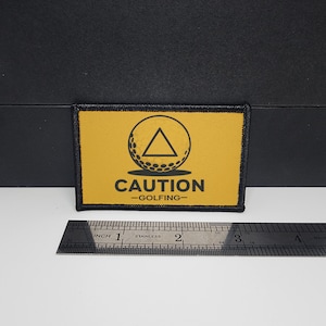 May include: A rectangular patch with a yellow background and black border. The patch features a golf ball inside a circle with a triangle above the word "CAUTION" and "-GOLFING-" below. A stainless steel ruler is next to the patch.