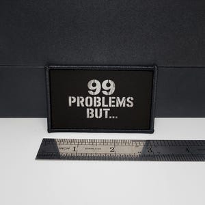 Può includere: Toppa rettangolare nera con la scritta "99 PROBLEMS BUT..." in bianco. La toppa misura circa 7,6 cm di lunghezza e 3,8 cm di larghezza. Un righello di metallo è posto sotto.