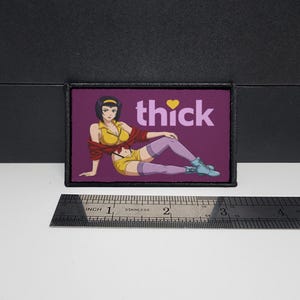 May include: A rectangular embroidered patch with a purple background featuring an anime character with short dark hair, wearing a yellow top and shorts, a red jacket, and purple stockings. The text thick with a yellow heart is printed above.