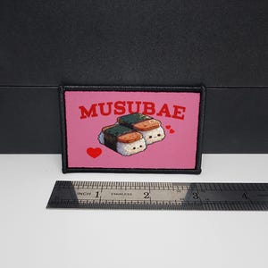 May include: A rectangular patch with a pink background features the text "MUSUBAE" in red and two cartoon musubi characters. The musubi have white rice, spam, and nori. Two red hearts are also present. The patch is approximately 2 inches wide.