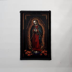 Peut inclure: Écusson rectangulaire avec une bordure noire, représentant une figure squelettique en position religieuse. La figure porte des robes rouges et bleu foncé, entourée d'un halo doré. Des fleurs oranges et un crâne sont en bas.