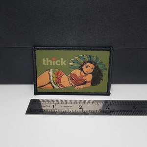 May include: A rectangular patch with a cartoon illustration of a woman in a tropical outfit, with the word "thick" above her. The patch is green with a black border. The woman is wearing a red top and a skirt with floral and feather accents.