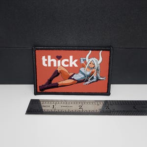 Puede incluir: Parche rectangular con fondo rojo y la palabra "thick" en blanco con un símbolo de corazón. El parche presenta una ilustración de estilo anime de una mujer con cabello blanco y cuernos. El parche mide aproximadamente 7,6 cm de ancho.
