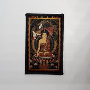 Pode incluir: Obra de arte retangular com uma figura de Buda sentado em um manto marrom, cercado por uma auréola dourada e figuras. A obra de arte tem uma borda trançada preta e está em um fundo preto com detalhes dourados. O Buda está sentado em uma flor de lótus.
