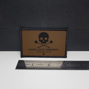 May include: A rectangular brown patch with black stitching and a black border. The patch features a black skull and crossbones design above the text "CAPS LOCK ORIGINAL COUNTRY CLUB". A stainless steel ruler is next to the patch.