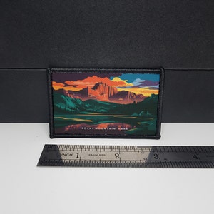 Könnte beinhalten: Rechteckiger Aufnäher mit schwarzem Rand, der eine Landschaft des Rocky Mountain Parks zeigt. Das Bild zeigt Berge, einen See und einen Sonnenuntergang. Der Text "ROCKY MOUNTAIN PARK" ist unten aufgedruckt.