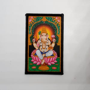 Könnte beinhalten: Ein rechteckiger Aufnäher mit einer lebendigen Illustration von Ganesha, der hinduistischen Gottheit. Ganesha ist auf einer rosa Lotusblume sitzend dargestellt, umgeben von einem grünen und orangefarbenen Bogen. Der Aufnäher hat einen schwarzen Rand.