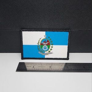 Könnte beinhalten: Ein rechteckiger Aufnäher mit der Flagge von Rio de Janeiro, Brasilien. Die Flagge hat einen blau-weißen Hintergrund mit dem Staatswappen in der Mitte. Ein Edelstahl-Lineal befindet sich darunter, mit Messungen in Zentimetern.