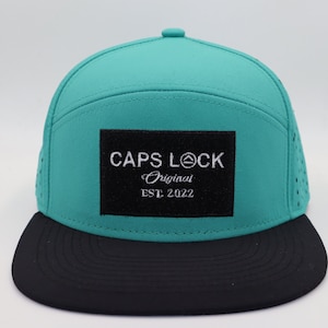 Puede incluir: Gorra de béisbol turquesa y negra con el texto "CAPS LOCK Original EST. 2022" en un parche cuadrado negro. La gorra tiene una visera plana y pequeños orificios de ventilación en los laterales.