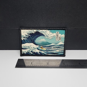 May include: Rectangular patch featuring a stylized wave design with a surfer and the text "Surfs Up." The patch is approximately 3 inches wide and 2 inches tall, with a black border. The design includes shades of blue, white, and a hint of yellow.