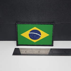 Puede incluir: Un parche rectangular con la bandera de Brasil. La bandera tiene un fondo verde con un rombo amarillo en el centro. Dentro del rombo hay un círculo azul con estrellas blancas y una pancarta que dice "Ordem e Progresso". El parche está bordeado con costuras negras.