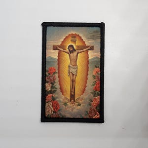 Könnte beinhalten: Rechteckiger Aufnäher mit einem religiösen Bild von Jesus Christus am Kreuz. Das Bild hat eine warme Farbpalette mit einem goldenen Heiligenschein um die Figur. Rote und rosa Rosen umrahmen den unteren Teil, und der Aufnäher hat einen schwarzen Rand.
