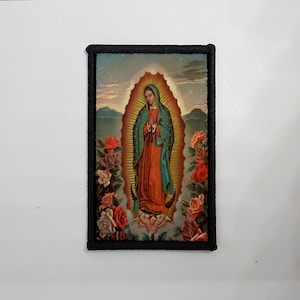May include: Rectangular patch featuring an illustration of Our Lady of Guadalupe. The image depicts a figure in a red and teal robe, surrounded by an oval of light and roses. The patch has a black border and is set against a white background.