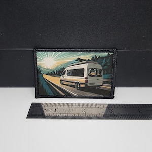 May include: Rectangular patch featuring a white camper van driving on a road towards a bright sun. The background includes mountains and trees. The patch has a black border and measures approximately 3 inches wide.