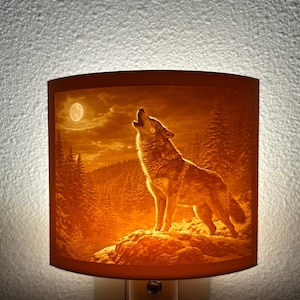 May include: A night light with a warm orange glow, featuring a detailed image of a howling wolf under a full moon. The scene includes a forest silhouette, creating a serene, nature-inspired ambiance. The night light is designed to plug into a standard wall outlet.