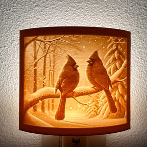 May include: A nightlight with a 3D carved image of two red cardinals perched on a branch in a snowy forest. The light casts a warm glow through the carved design.