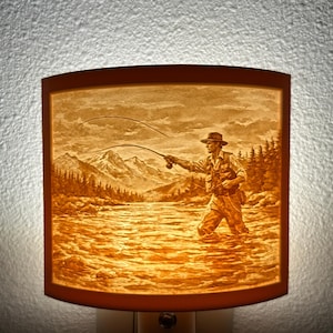 May include: A rectangular night light with a warm, amber glow. The image depicts a fly fisherman in a river with mountains in the background. The fisherman is wearing a hat and waders, casting a line. The scene is detailed and evokes a sense of nature and outdoor recreation.