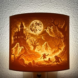 May include: A Halloween-themed night light with a spooky scene. The image depicts a haunted house, ghosts, goblins, and a full moon. The color palette is primarily orange and brown, creating a warm, eerie glow.