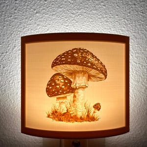 May include: A nightlight with a white ceramic shade featuring a detailed illustration of three mushrooms with brown caps and white stems. The mushrooms are surrounded by green grass and leaves.