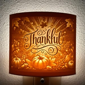 May include: A nightlight with a cream-colored shade featuring a carved design of fall leaves, pumpkins, and the word "Thankful".