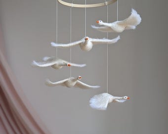 May include: A white felt mobile with five geese flying in a circle. The geese are suspended from a wooden ring.