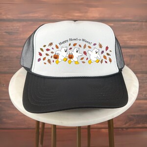 May include: A black and white trucker hat with a white front panel featuring the text "Happy Howl-o-Ween!" and a graphic of three cartoon ghosts carrying jack-o'-lanterns. The design includes autumn leaves. The hat has a mesh back.