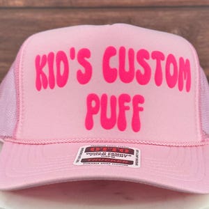May include: Pink trucker hat with a pink mesh back. The hat has the words "Kid's Custom Puff" in a pink bubble font.