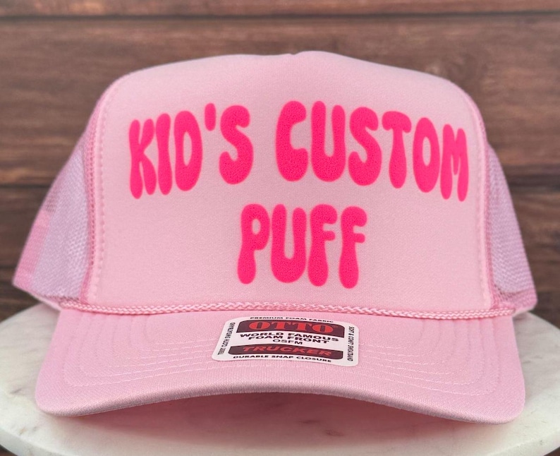 May include: Pink trucker hat with a pink mesh back. The hat has the words "Kid's Custom Puff" in a pink bubble font.