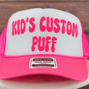 May include: Pink trucker hat with a white front panel. The front panel has the text "Kid's Custom Puff" in pink bubble letters. The hat has a black and white label with the text "OTTO World Famous Foam Front 1SFM TRUCKER DURABLE SNAP CLOSURE".