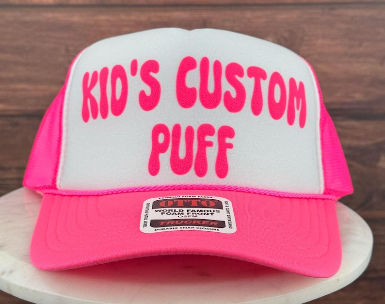 May include: Pink trucker hat with a white front panel. The front panel has the text "Kid's Custom Puff" in pink bubble letters. The hat has a black and white label with the text "OTTO World Famous Foam Front 1SFM TRUCKER DURABLE SNAP CLOSURE".