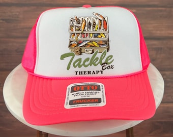 Puede incluir: Gorra de camionero con un panel frontal blanco y una visera y una parte trasera de malla de color rosa brillante. La parte delantera presenta un gráfico de una caja de aparejos con las palabras "Tackle Therapy".
