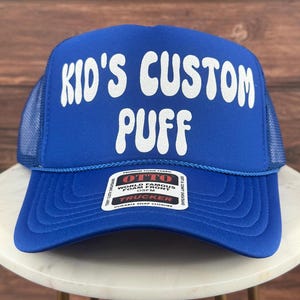 May include: A blue trucker hat with a white "Kid's Custom Puff" logo. The hat has a mesh back and a snap closure.