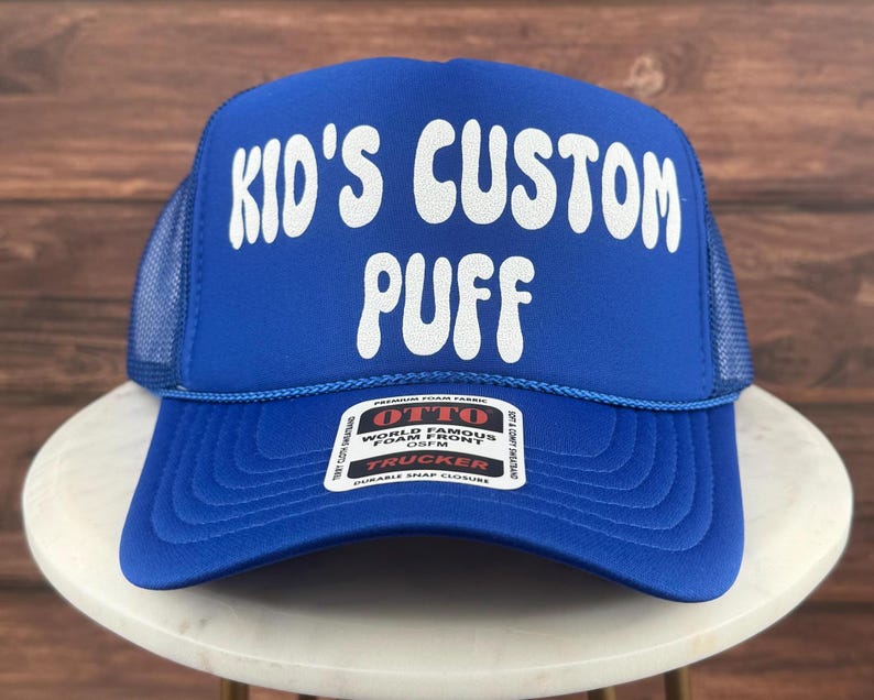 May include: A blue trucker hat with a white "Kid's Custom Puff" logo. The hat has a mesh back and a snap closure.