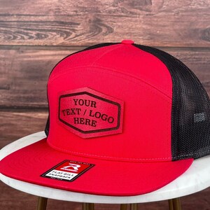 Op de afbeelding: Een rode en zwarte truckercap met een platte klep en een bruin leren patch met de tekst "YOUR TEXT/LOGO HERE".