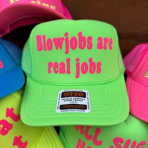 May include: A selection of trucker caps in vibrant neon shades. The main cap is a vivid green, featuring the pink text "Blowjobs are real jobs". Other caps display phrases such as "Hurting for a ring" and "All no sugar daddy".