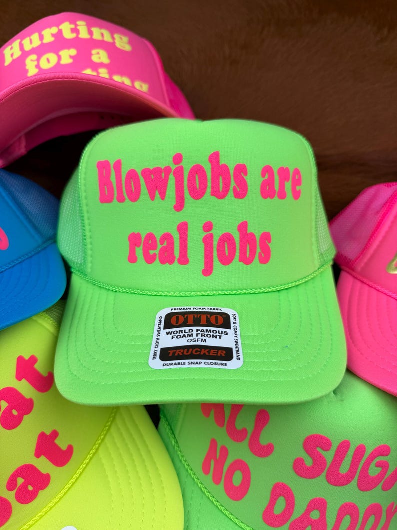 May include: A selection of trucker caps in vibrant neon shades. The main cap is a vivid green, featuring the pink text "Blowjobs are real jobs". Other caps display phrases such as "Hurting for a ring" and "All no sugar daddy".