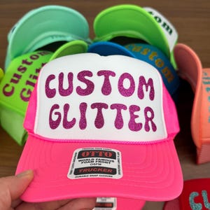 May include: A pink trucker hat with a white front panel featuring the text "CUSTOM GLITTER" in glittery magenta letters. The hat has a mesh back and a curved brim. Other hats in various colors are visible in the background.