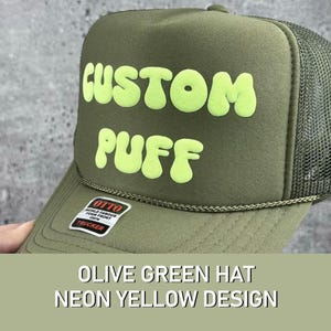 May include: Olive green trucker hat with a neon yellow "CUSTOM PUFF" design. The hat has a mesh back and a black label that says "OTTO WORLD FAMOUS FOAM FRONT CAP TRUCKER".