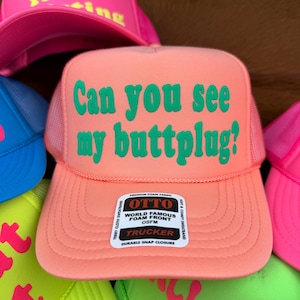 May include: A peach-coloured trucker hat with the text "Can you see my buttplug?" in green. The hat has a mesh back and a snap closure. Other hats with neon text are visible.