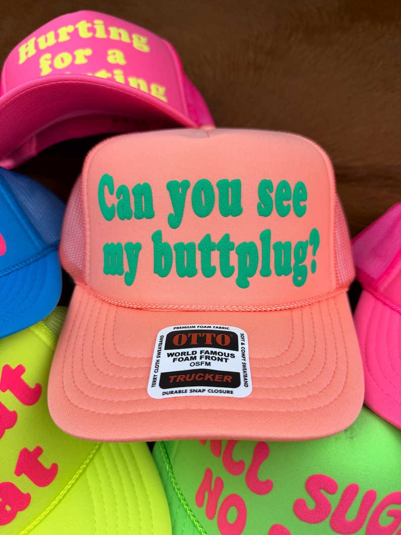 May include: A peach-coloured trucker hat with the text "Can you see my buttplug?" in green. The hat has a mesh back and a snap closure. Other hats with neon text are visible.